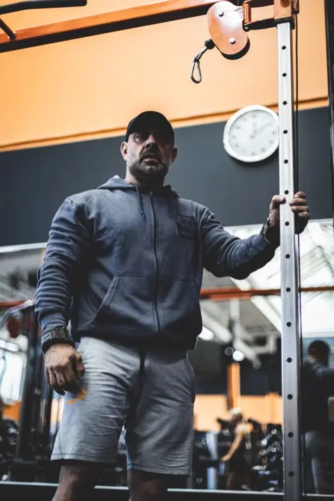 Ashkan Aghili står vid ett kabelställ på gymgolvet med handen på stativet